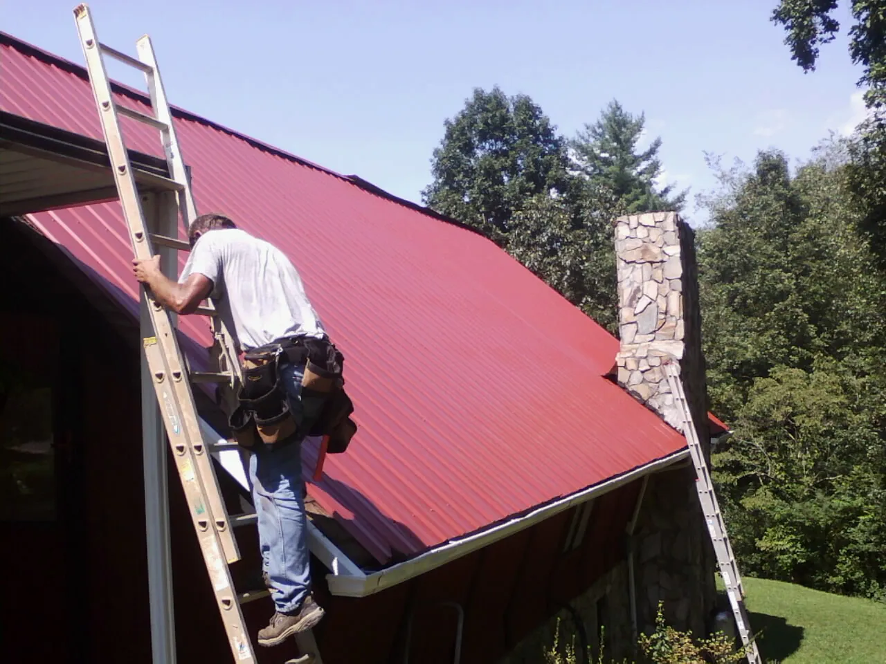 Roofer on a ladder beside a red metal roof and stone chimney — Soffit & Fascia Repair by Roofing Brothers in Herculaneum